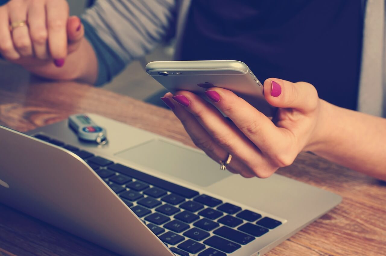 Osoba siedzi przed laptopem i w ręku trzyma telefon
