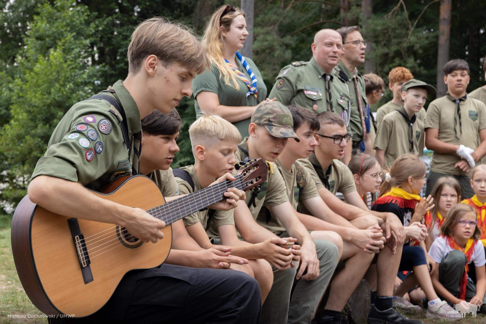 Harcerze śpiewają a jeden z nich gra na gitarze