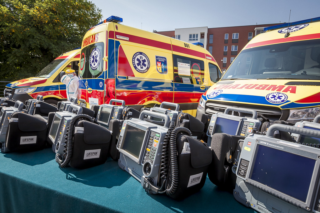Na pierwszym planie na stole leżą defibrylatory, w tle stoją trzy ambulanse.